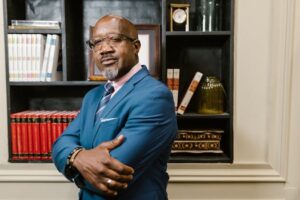African American man in blue suit with arms crossed, standing confidently in a library.